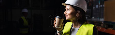 Téléchargez les photos : Bannière de femme heureuse en chapeau dur tenant tasse en papier, employé de l'entrepôt pendant la pause café - en image libre de droit