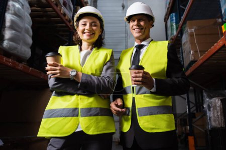 Cheerful logistics team with hard hats and coffee smiling during a break in warehouse, man and woman