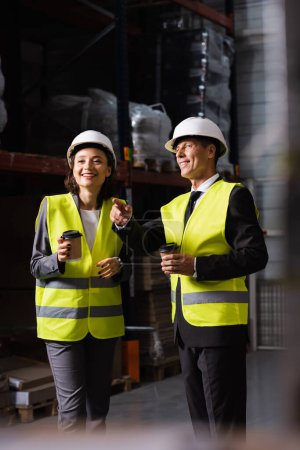 equipo logístico, feliz supervisor en sombrero duro y café mostrando algo a un colega en el almacén