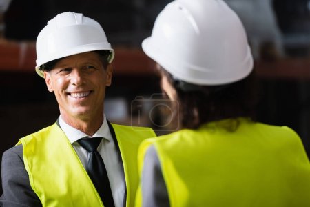 happy middle aged supervisor looking at his female colleague in hard hat on blurred foreground