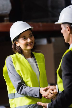 supervisor and his happy female colleague in hard hat and safety vest shaking hands in warehouse