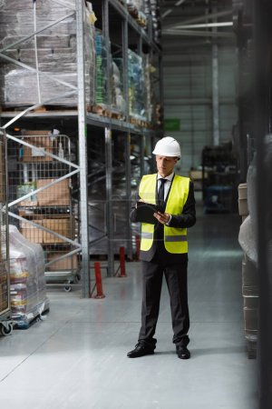 confident middle aged supervisor in safety vest and hard hat writing on clipboard in warehouse