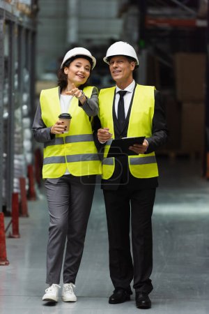 happy logistics workers with hard hats walking with coffee near inventory while inspecting warehouse