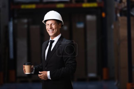 heureux homme d'affaires d'âge moyen en casque dur et costume tenant café dans l'entrepôt, professionnel