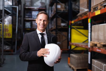 happy middle aged businessman in suit holding hard hat in warehouse, professional headshot