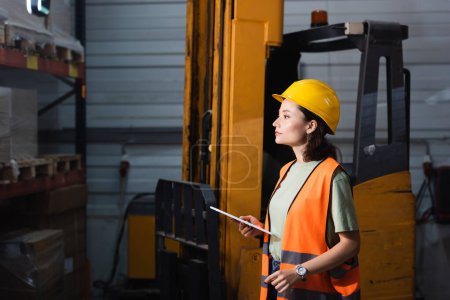 female warehouse worker in hard hat and safety vest holding digital tablet near forklift, cargo