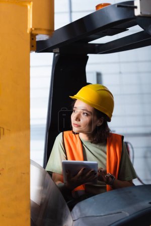 Foto de Trabajadora de almacén pensativa en sombrero duro y chaleco de seguridad usando tableta y sentada en carretilla elevadora - Imagen libre de derechos