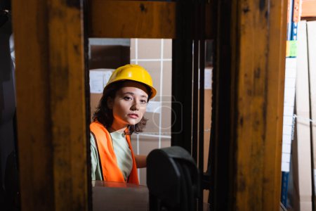female forklift operator in hard hat and safety vest looking at camera in warehouse, cargo