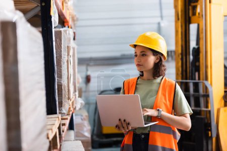 trabajadora con sombrero duro y chaleco de seguridad usando laptop mientras revisa inventario en almacén