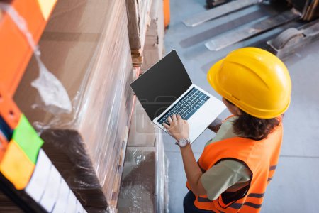 female worker in hard hat and safety vest using laptop while checking cargo in warehouse, top view
