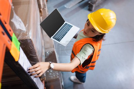 female warehouse worker in hard hat and safety vest using laptop while checking cargo, top view