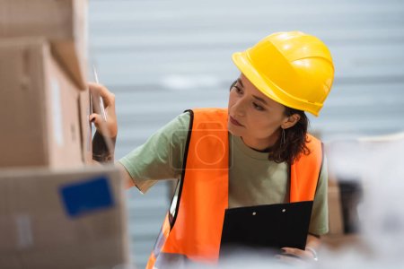 female warehouse worker in safety vest holding clipboard while checking cargo, tracking system