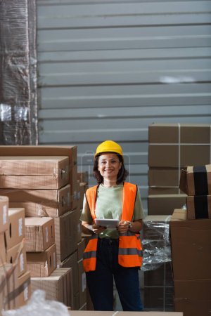 cheerful female warehouse worker in safety vest and hard hat holding digital tablet near cargo