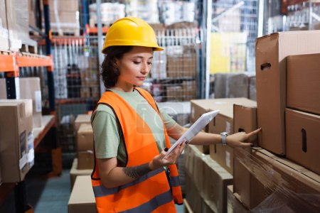 female warehouse worker in safety vest and hard hat holding digital tablet and inspecting cargo