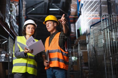Two warehouse workers discussing logistics, middle aged man pointing near woman with folder