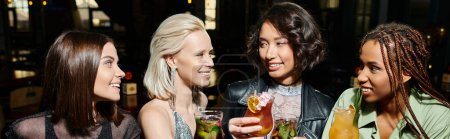 smiling multiethnic women holding cocktail glasses while spending time in bar, horizontal banner