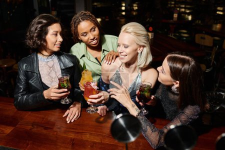 novias multiétnicas sonrientes y elegantes con copas de cóctel hablando en el bar, ocio nocturno