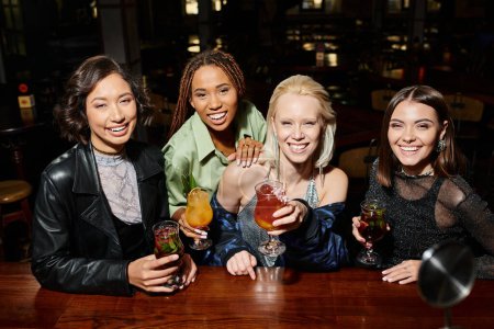Téléchargez les photos : Charmante et heureuse copines multiethniques avec des verres à cocktail regardant la caméra dans le bar - en image libre de droit