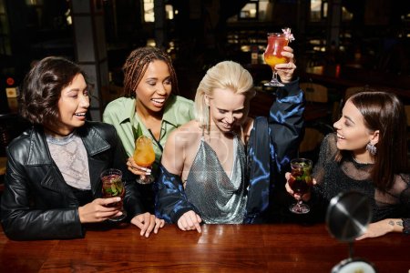 excited blonde woman toasting with cocktail glass near elegant multiethnic girlfriends in bar