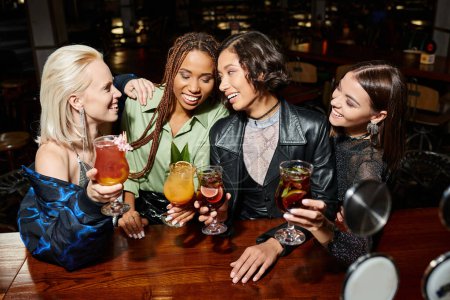 verführerische und elegante multiethnische Freundinnen, die mit Cocktails anstoßen, während sie sich in der Bar ausruhen, im Nachtleben