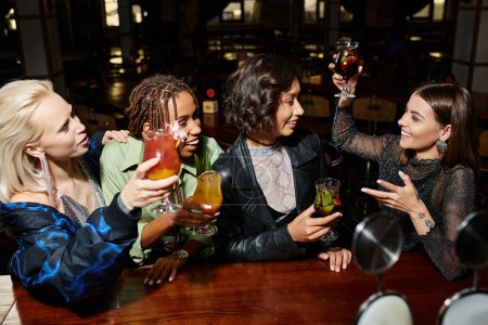 cheerful woman toasting with cocktail near multiracial girlfriends in nigh bar, vibrant lifestyle