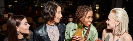 happy and trendy multiracial girlfriends with cocktail glasses talking in bar, horizontal banner