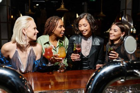 fröhliche multikulturelle Freundinnen mit Cocktailgläsern, die während des Gesprächs in der Bar lächeln, Polterabend