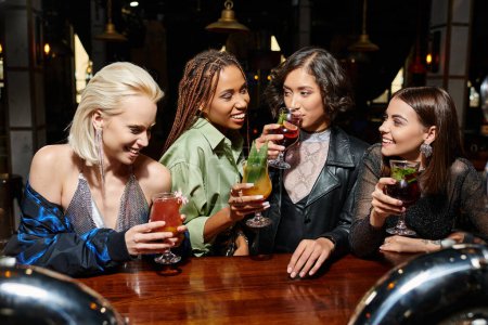 young asian woman drinking cocktail near cheerful multiethnic girlfriends in bar, party time