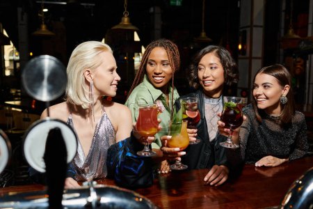 Téléchargez les photos : Copines multiculturelles insouciantes griller avec des cocktails dans une atmosphère vibrante de bar de nuit - en image libre de droit