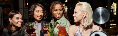 des amies multiculturelles souriantes et élégantes tenant des verres à cocktails dans un bar, bannière horizontale
