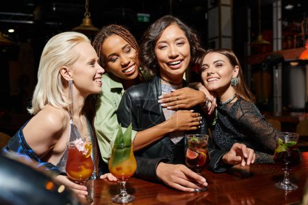 happy african american woman embracing asian girlfriend in cocktail bar, hen party at nighttime