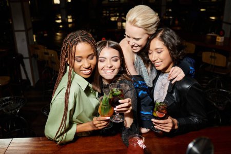 joyful multicultural girlfriends clinking cocktail glasses and smiling with closed eyes in bar