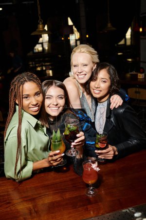 smiling multiethnic girlfriends with cocktail glasses embracing and looking at camera in bar