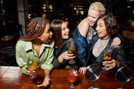copines multiethniques excitées et branchées avec des verres à cocktail parlant au bar, atmosphère vibrante