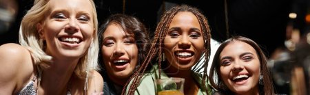groupe diversifié de copines multiethniques souriant à la caméra dans le bar de nuit, bannière horizontale