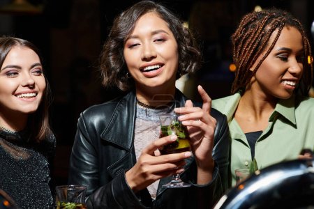 junge und trendige Asiatin mit Cocktailgläsern, die neben multiethnischen Freundinnen in der Nachtbar lächelt
