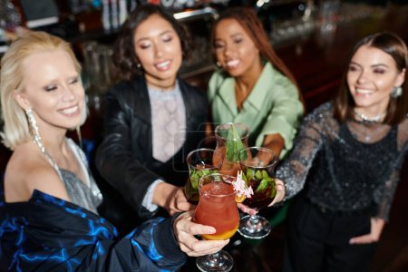 lächelnde und elegante multiethnische Freundinnen, die in einer Bar vor verschwommenem Hintergrund Cocktails klappern