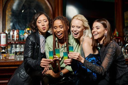 Photo for Joyful and stylish multiracial girlfriends clinking shot glasses during festive party in bar - Royalty Free Image