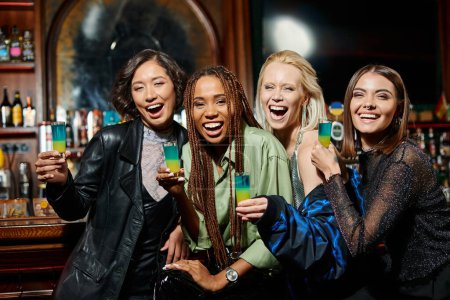 Photo for Glamorous multiracial girlfriends with shot glasses laughing and looking at camera in bar, party - Royalty Free Image