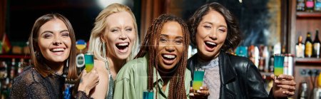Photo for Stylish multiracial girlfriends with shot glasses laughing and looking at camera in bar, banner - Royalty Free Image