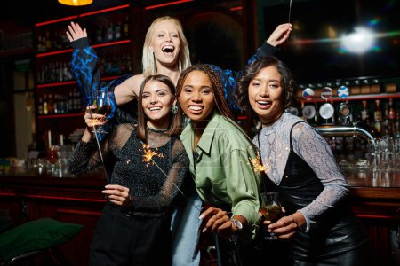 Foto de Alegres novias multiétnicas con cócteles y chispas festivas divirtiéndose en el bar, fiesta festiva - Imagen libre de derechos