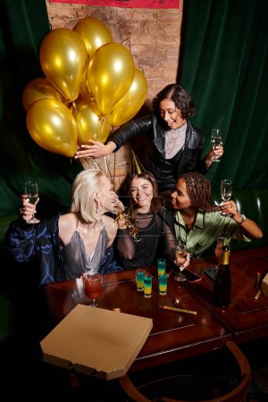 glamorous multiethnic women celebrating birthday with alcohol drinks and golden balloons in bar