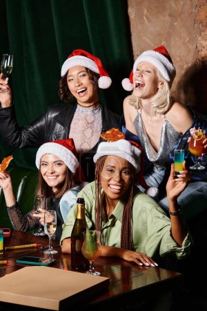 mujeres multiétnicas jóvenes y despreocupadas en sombreros de Navidad celebrando el Año Nuevo en el bar, fiesta glamorosa