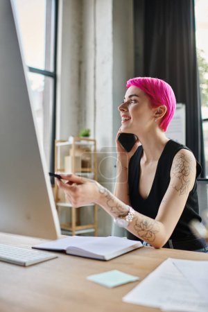 alegre mujer de negocios tatuada en traje casual hablando por teléfono mientras trabaja duro en la computadora