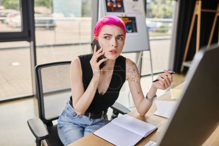 focused pink haired businesswoman in urban attire talking by phone while working hard at computer