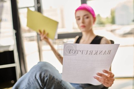 verschwommene junge Frau mit rosa Haaren in lässiger Kleidung liest Dokument mit dem Wort Bankrott darauf