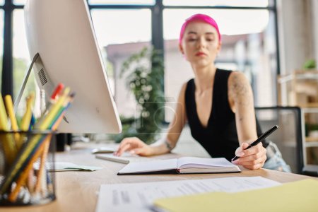 blurred photo of pink haired businesswoman taking notes while working hard, focus on her computer