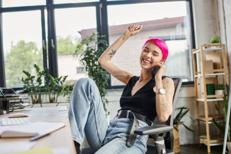 Téléchargez les photos : Joyeuse femme d'affaires aux cheveux roses avec des tatouages en tenue urbaine décontractée parlant par téléphone au bureau, - en image libre de droit