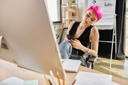 joven mujer trabajadora de pelo rosa con tatuajes mirando su teléfono móvil mientras trabaja en la oficina