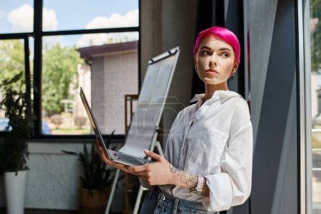 pensive pink haired woman with tattoos holding laptop and looking away at office, business concept